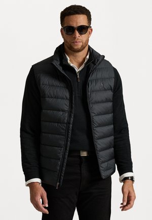 Schwarze Steppweste mit horizontalem Quilt-Design, hohem Kragen, Reißverschluss, getragen über einem schwarzen Pullover, kombiniert mit dunklen Jeans und Sonnenbrille.