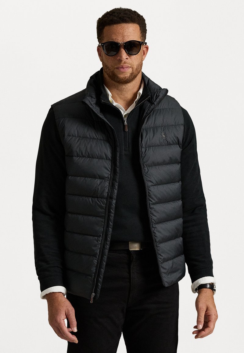 Gilet matelassé noir avec un design quilté horizontal, col haut, fermeture éclair, porté sur un pull noir, associé à un jean sombre et des lunettes de soleil.