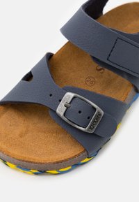 Sandalia de cuero azul marino con tira de velcro y hebilla de metal. Presenta una plantilla texturizada y una suela de patrón multicolor. Talla 25.