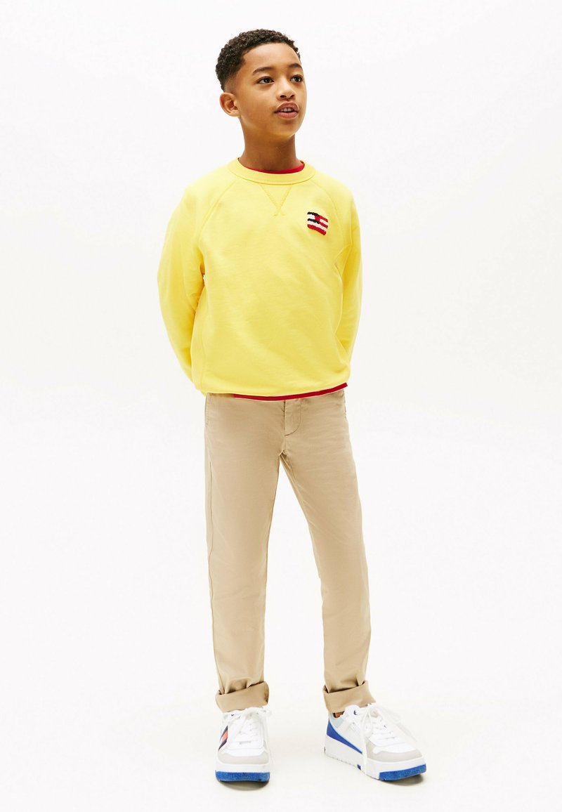 Felpa gialla in cotone con scollatura rotonda e rifiniture rosse. Pantaloni beige e sneakers bianche con accenti blu. Stilizzato in modo informale, in posizione in piedi.