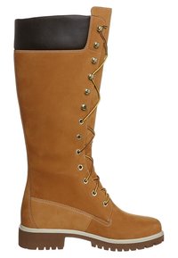 Bota de gamuza en tono marrón claro, de caña alta, con un puño de cuero marrón, herrajes dorados y un diseño de cordones con ojales hexagonales. Suela de goma.