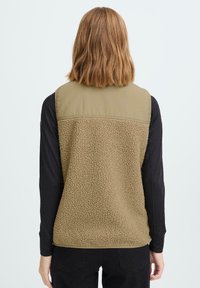 Fleece-Weste in Khaki mit strukturiertem Rücken und glattem Stoff an den Schultern. Getragen über einem schwarzen Langarmshirt mit lässiger Passform.