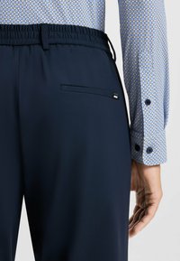 Pantaloni blu navy con cintura elastica, dotati di una tasca posteriore e una discreta etichetta con logo. La camicia con polsini presenta un design a motivo geometrico.