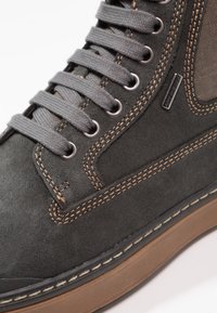 Zapatilla de ante gris con cordones grises, costuras marrones, suela de goma y panel de malla lateral. Etiqueta de marca destacada en el lateral.