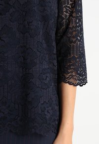 Top in pizzo blu navy con motivi floreali, maniche a tre quarti, orlo smerlato e trasparenza su una base solida.