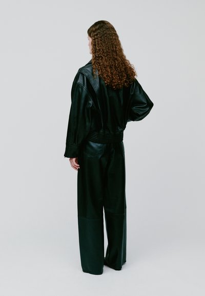 Žena s dugom kovrčavom kosom, odjevena u crnu kožnu jaknu i široke crne kožne hlače, stoji okrenuta leđima na jednobojnoj svijetlosivoj pozadini.