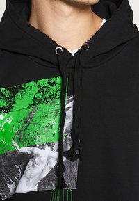 Sweat à capuche noir avec un graphisme aux accents verts et noirs, cordons de serrage, bords usés, et une image imprimée sur le devant.