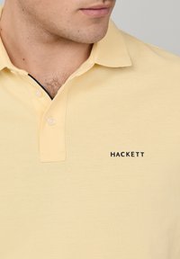 Homme portant un polo jaune pâle avec col et logo noir "HACKETT" sur la poitrine gauche, montrant partiellement le cou et le menton.