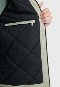 Das Innere der gesteppte Jacke ist schwarz mit einem hellgrünen Besatz. Sie verfügt über eine Reißverschlusstasche auf der linken Seite. Der Stoff hat eine glatte, leicht glänzende Textur.