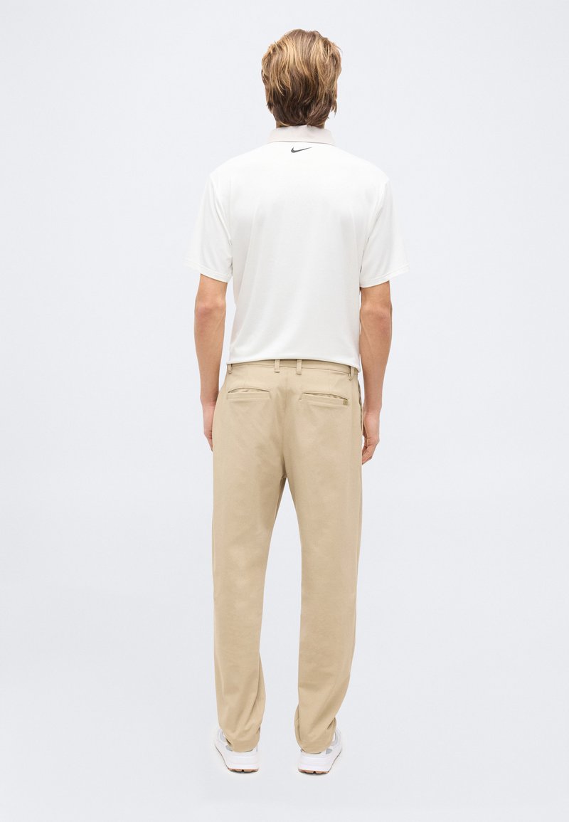 Homme debout, vu de dos, portant un polo Nike blanc, un pantalon beige et des baskets blanches sur un fond clair uni.