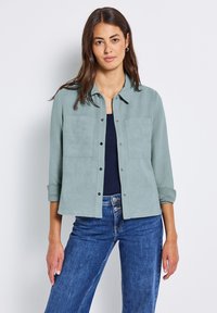 Žena s dlouhými hnědými vlasy má na sobě světle modrou košili s knoflíky přes námořnickou blůzu a modré džíny, stojí na pozadí bílé stěny.