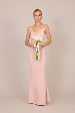 Kvinde i en gulvlængde blegrosa kjole, der holder en hvid calla-lilje buket bundet med et hvidt bånd mod en ensfarvet beige baggrund.