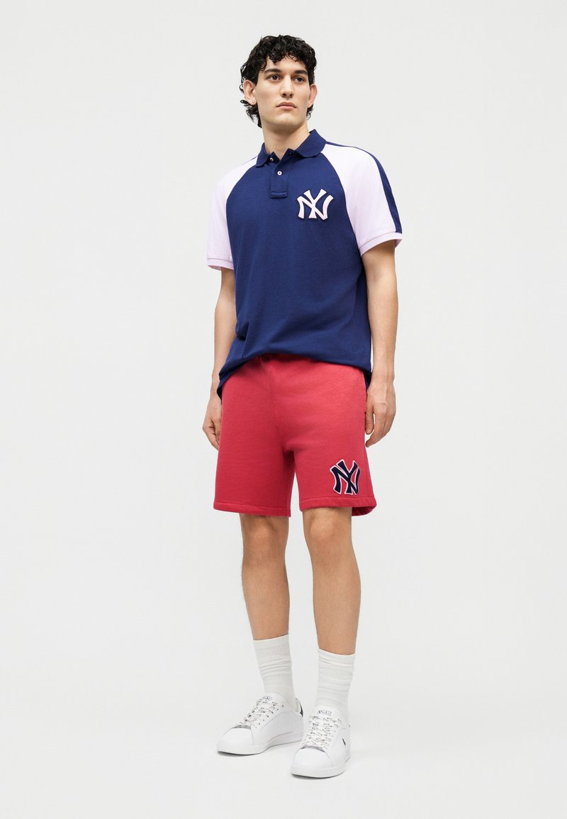 Jonge man die een navyblauw-witte New York Yankees poloshirt draagt, met rode korte broek, witte sokken en witte sneakers, staand tegen een effen achtergrond.