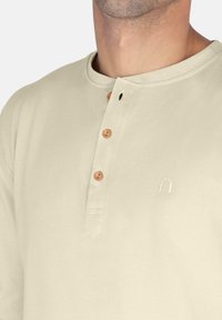 Beige Henley-Shirt mit drei Holzknöpfen, Rundhalsausschnitt und kurzer Knopfleiste. Auf der Brust ist ein minimalistisches Logo gestickt.