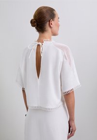 Haut blanc à manches courtes avec une ouverture en forme de clé dans le dos, featuring un tissu texturé et une bordure en dentelle le long des manches et de l'ourlet.