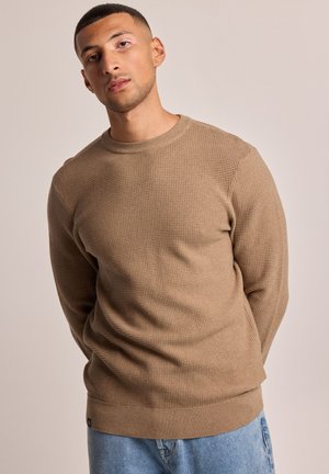Maglione beige lavorato a maglia con motivo a rilievo, scollo rotondo e polsini a costine. Abbinato a jeans di denim azzurro chiaro.
