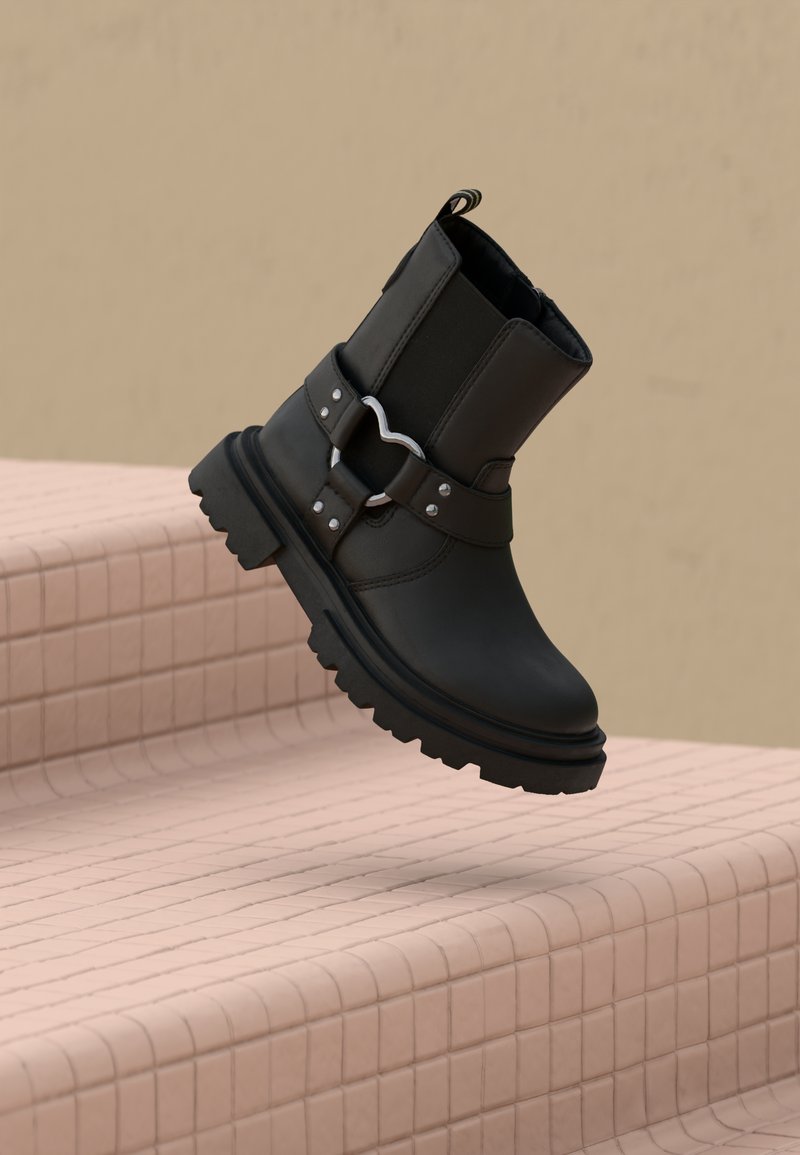 Sort ankelstøvle med læder- og elastikdesign, struktureret gummisål, sølvhardwaredetalje og en hævet slæb ved bagsiden.