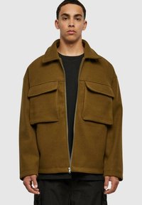 Veste zippée marron en tissu doux, dotée de deux grandes poches avant, d'un col rabattu et d'une coupe décontractée. Portée par-dessus une chemise noire.