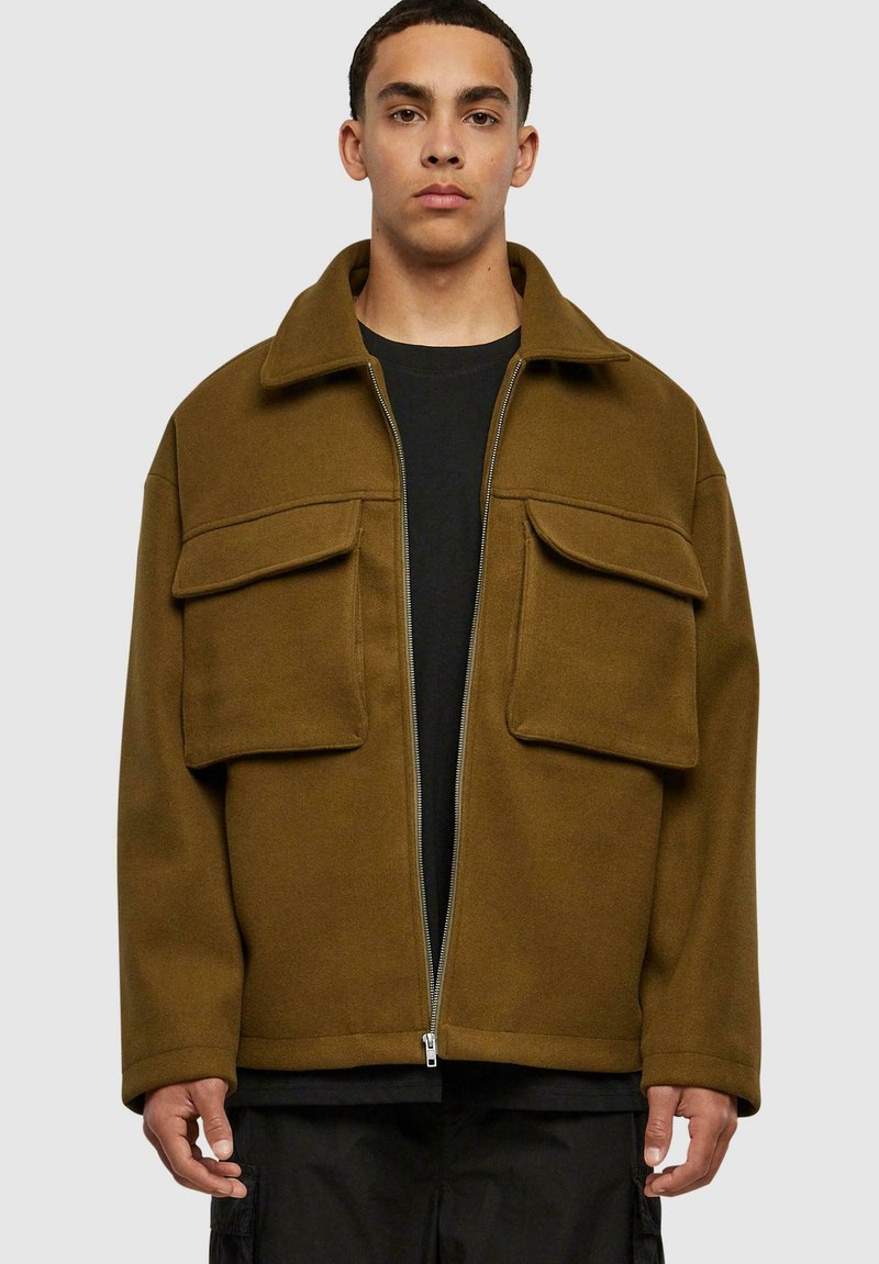 Veste zippée marron en tissu doux, dotée de deux grandes poches avant, d'un col rabattu et d'une coupe décontractée. Portée par-dessus une chemise noire.