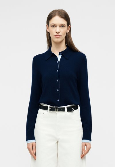 Femme aux longs cheveux bruns portant un cardigan bleu marine boutonné sur un haut bleu clair, un pantalon blanc et une ceinture noire, debout devant un fond blanc.