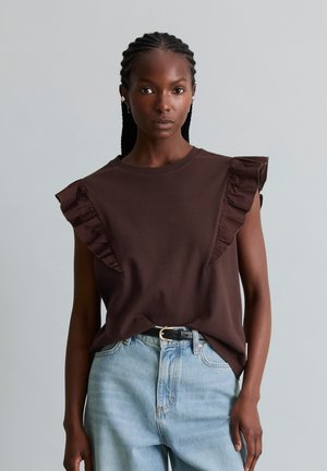 Femme aux cheveux tressés portant un haut marron sans manches avec des volants et un jean bleu clair avec une ceinture noire, debout devant un fond gris uni.