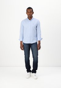 Camicia a quadri blu e bianchi con colletto button-down, abbinata a jeans scuri e sneakers bianche. Il modello si trova di fronte a uno sfondo bianco uniforme.