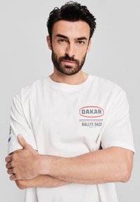 Biała bawełniana koszulka z grafiką "DAKAR RALLYE RACE" w kolorze czerwonym i niebieskim. Krótkie rękawy i okrągły dekolt w luźnym kroju.