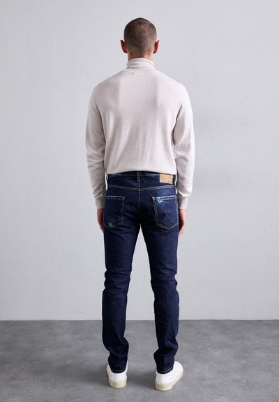 Mann, der mit dem Rücken zur Kamera steht, trägt einen beigen Rollkragenpullover, dunkelblaue Jeans mit leichter Abnutzung und weiße Sneaker vor einem grauen Boden und einer grauen Wand.
