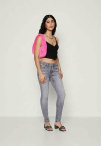 Top cropped preto com alças ajustáveis, jeans skinny de lavagem clara e uma bolsa de pelúcia rosa vibrante. Sandálias de salto pretas completam o looks.
