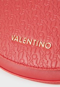 Röd texturerad läderväska med präglat "VALENTINO"-mönster och en metallisk guldlogotyp framtill, med släta kanter och sömdetaljer.