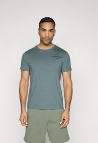 T-shirt leggera color verde acqua a girocollo con maniche corte, caratterizzata da un piccolo logo nero. Materiale in cotone morbido, vestibilità casual, abbinata a pantaloni corti verdi.