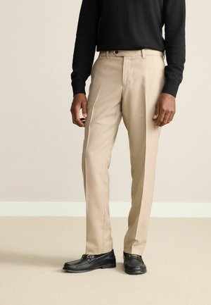 Beige Kleidungs-Hosen aus glattem Stoff, mit tailliertem Schnitt und flachem Bund. Getragen mit einem schwarzen Langarm-Shirt und schwarzen Slippern.