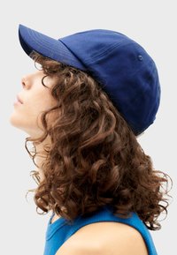 Marineblauwe katoenen baseballcap met een gebogen klep, zes-panelen constructie en zichtbare stiksels, gepositioneerd op een model met krullend haar.