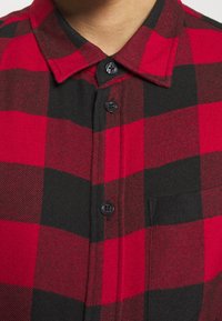 Gros plan sur une chemise en flanelle à carreaux rouge et noir avec des boutons noirs et un col, portée par une personne.
