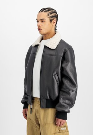 Chaqueta bomber de cuero negra con un suave cuello de borreguito blanco, puños acanalados y cierre de cremallera, llevada sobre una camisa de color claro y pantalones caqui.