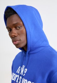 Felpa blu in pile con cappuccio regolabile, caratterizzata da un design del testo bianco "Nike Sportswear". Texture morbida e vestibilità rilassata.