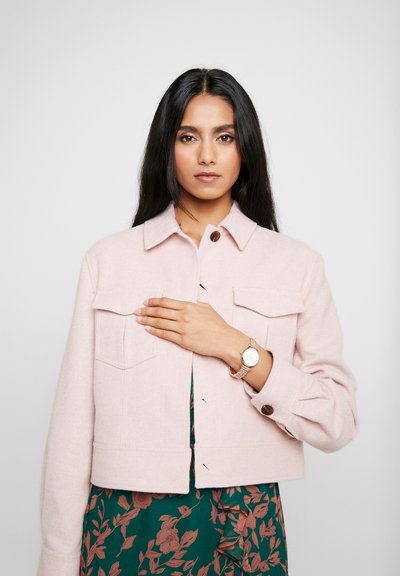 Femme aux longs cheveux noirs portant une veste rose pâle par-dessus une robe fleurie verte, la main sur la poitrine, portant une montre en or.
