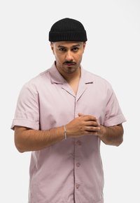 Camisa de manga corta de color rosa claro con cuello, con botones en la parte delantera y mangas enrolladas. Gorro de canalé negro y pulsera de plata en la muñeca.