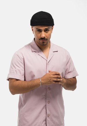 Lichtroze overhemd met korte mouwen, kraag, knoopsluiting aan de voorkant en omgeslagen mouwen. Zwarte geribde beanie en een zilveren armband om de pols.