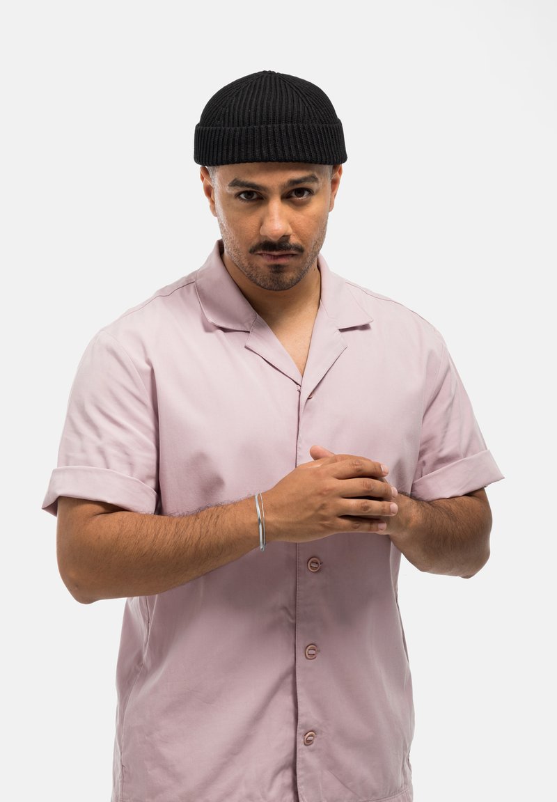 Camisa de manga corta de color rosa claro con cuello, con botones en la parte delantera y mangas enrolladas. Gorro de canalé negro y pulsera de plata en la muñeca.