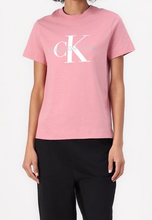 Vrouw draagt een roze Calvin Klein Jeans t-shirt met korte mouwen en een groot wit "ck" logo, gecombineerd met een zwarte broek.