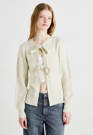 Cardigan lavorato a maglia di un delicato crema chiaro con bordi a costine, chiusura con fiocco, apertura frontale e maniche lunghe. Indossato sopra un top corto bianco e jeans di denim.