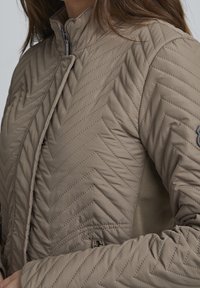 Jasnobrązowa pikowana kurtka z wzorem w jodełkę, zamkiem błyskawicznym i miękką tkaniną. Projekt zawiera boczną kieszeń z delikatnym akcentem.