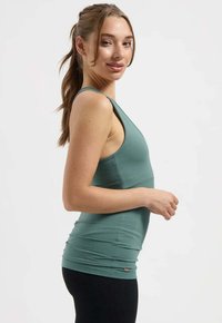 Tanktop van gedempt groen stof, aansluitend ontwerp, brede bandjes, geribbelde textuur en subtiele logo-accent bij de zoom. Gestyled met zwarte legging.