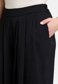 Zwarte broeken met een brede, elastische tailleband en zijzakken. Beschikt over een geplooid ontwerp en een zachte, gestructureerde stof.