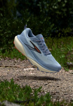 Hellblauer Brooks-Laufschuh mit dicker weißer Sohle, orangefarbenem Logo, schwebt über einem Schotterweg mit grünem Laub im Hintergrund.