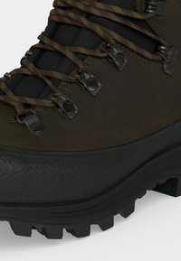 Close-up van een bruine wandelschoen met metalen oogjes, bruine patroonveters, een gewatteerde tong en een dikke zwarte zool met diepe groeven.