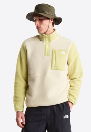 Sudadera de forro polar en crema y verde claro, con cuello de media cremallera, bolsillo con cremallera y logo de The North Face en el pecho. Acompañada de un sombrero tipo bucket en color oliva.