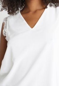 Haut blanc avec un col en V et des détails en dentelle aux épaules. Le tissu semble lisse avec un léger éclat.