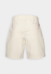 Shorts beige jusqu'aux genoux avec taille élastique, deux poches arrière boutonnées et une petite étiquette rouge sur la poche droite.